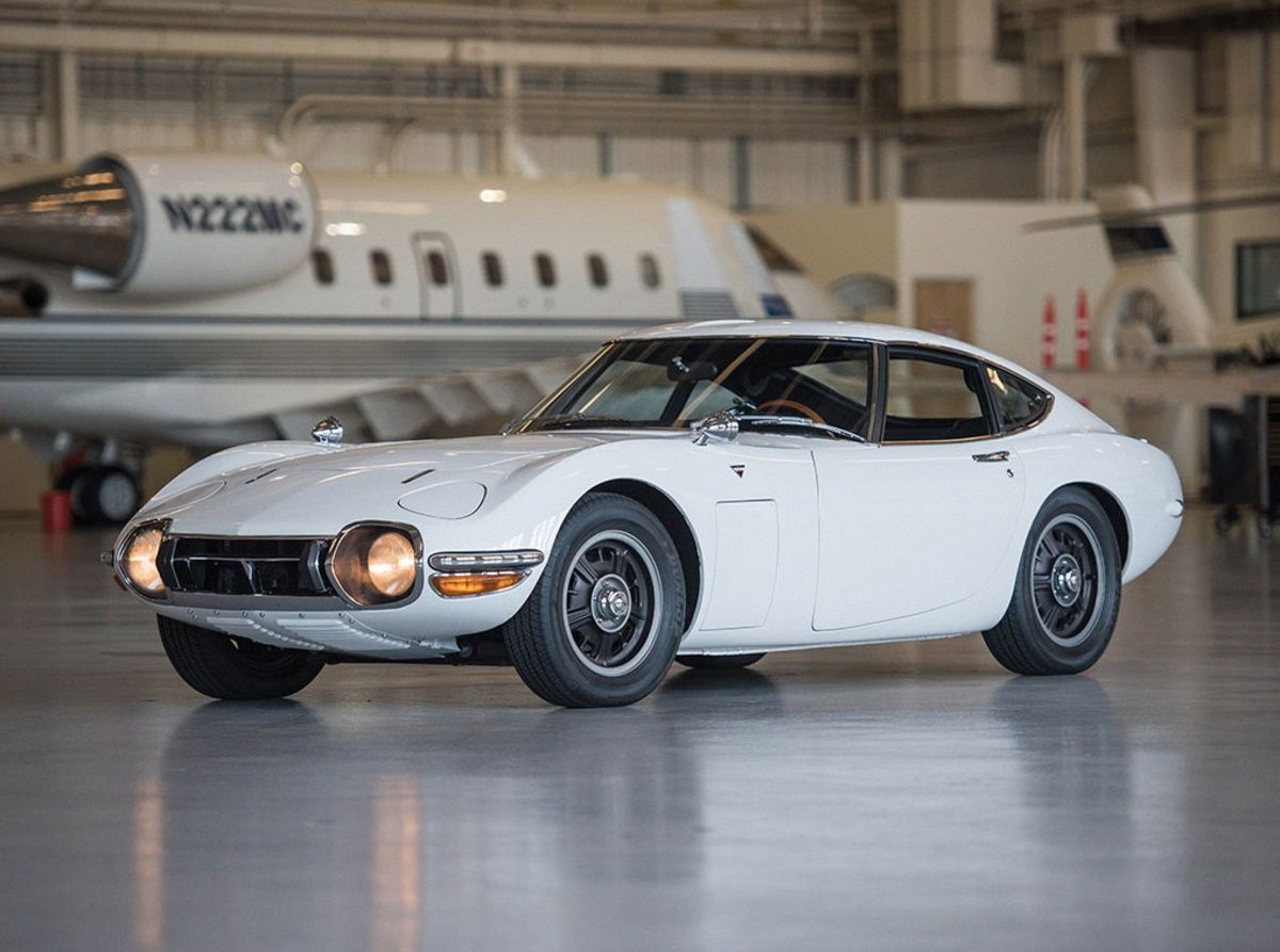 White Toyota 2000GT with its daylights on inside an aircraft hanger