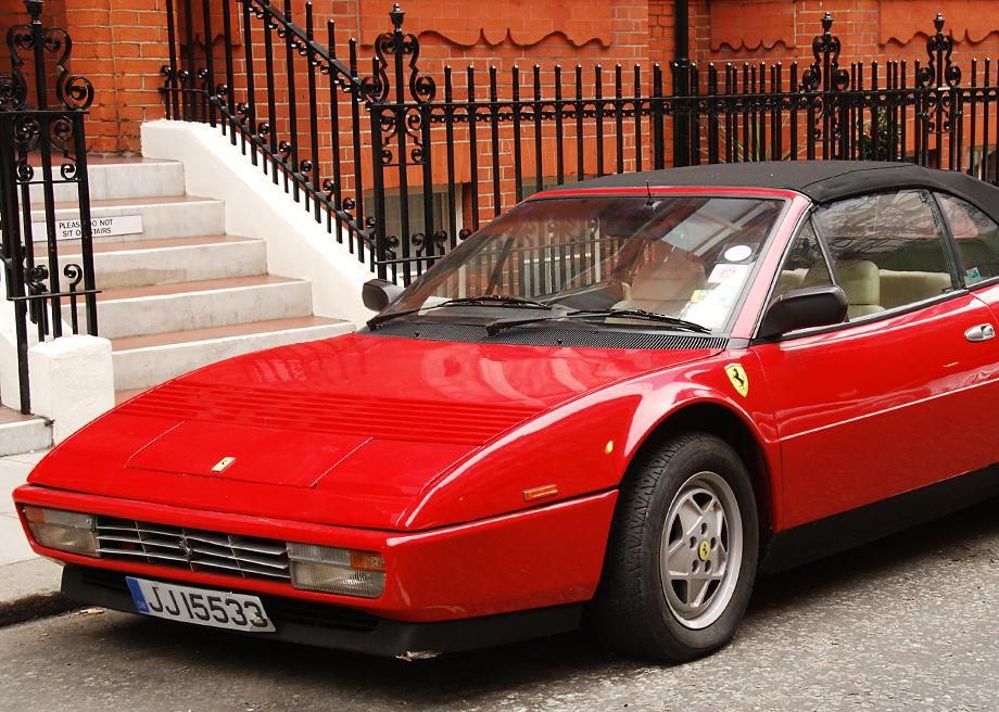 A 1985 Ferrari 3.2 Mondial Cabriolet with pop-up headlights stands parked on a city street.