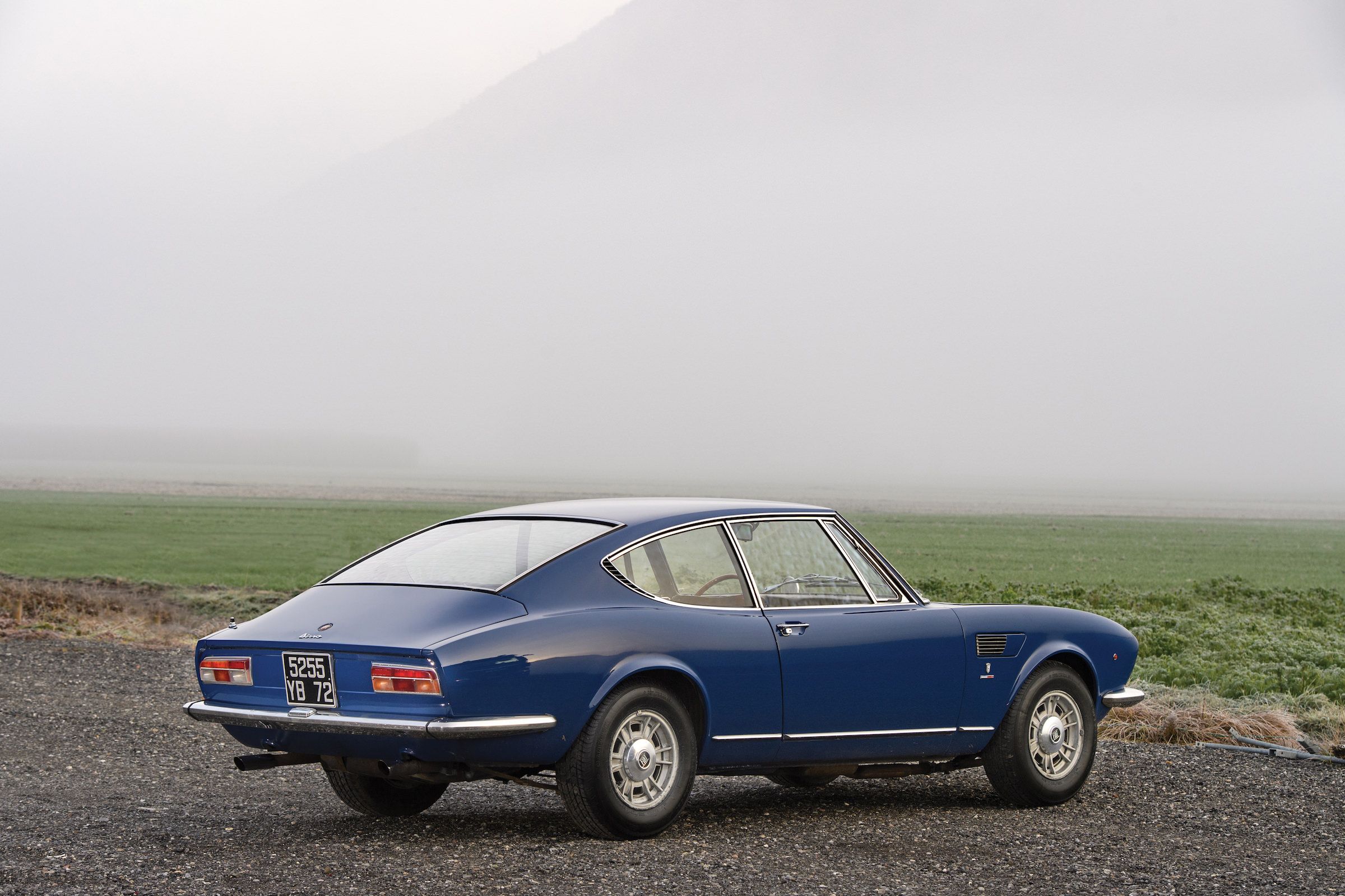 A blue Fiat Dino coupe stands parked on a foggy mountain road.
