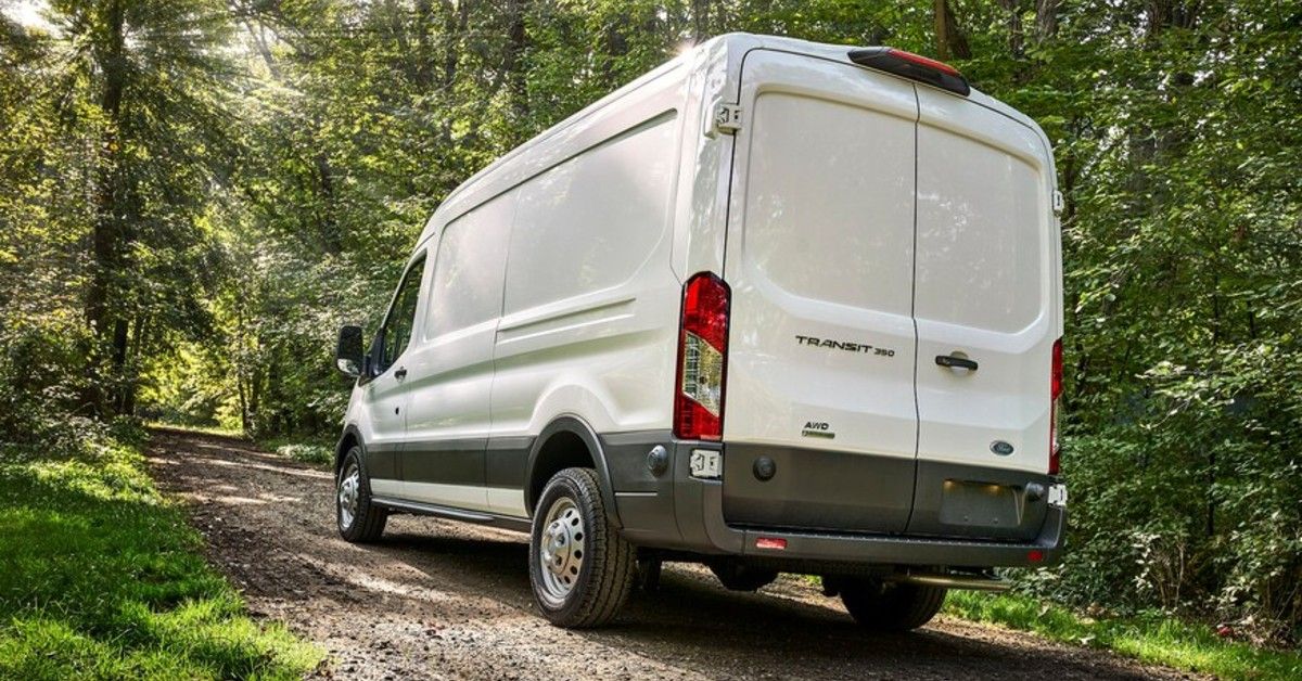 A white 2021 Ford Transit van drives through a forest on a clear day.