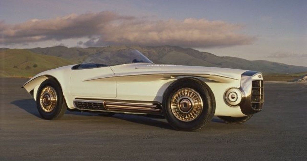 A 1965 Mercer Cobra Roadster stands parked on a road near mountains.