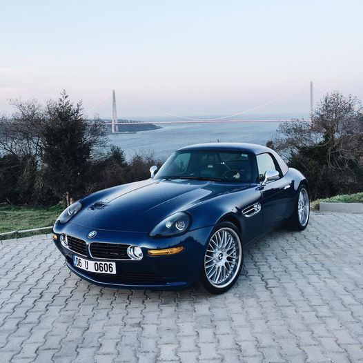 A blue BMW Z8 standing on a pavement with a water body and trees in the background.