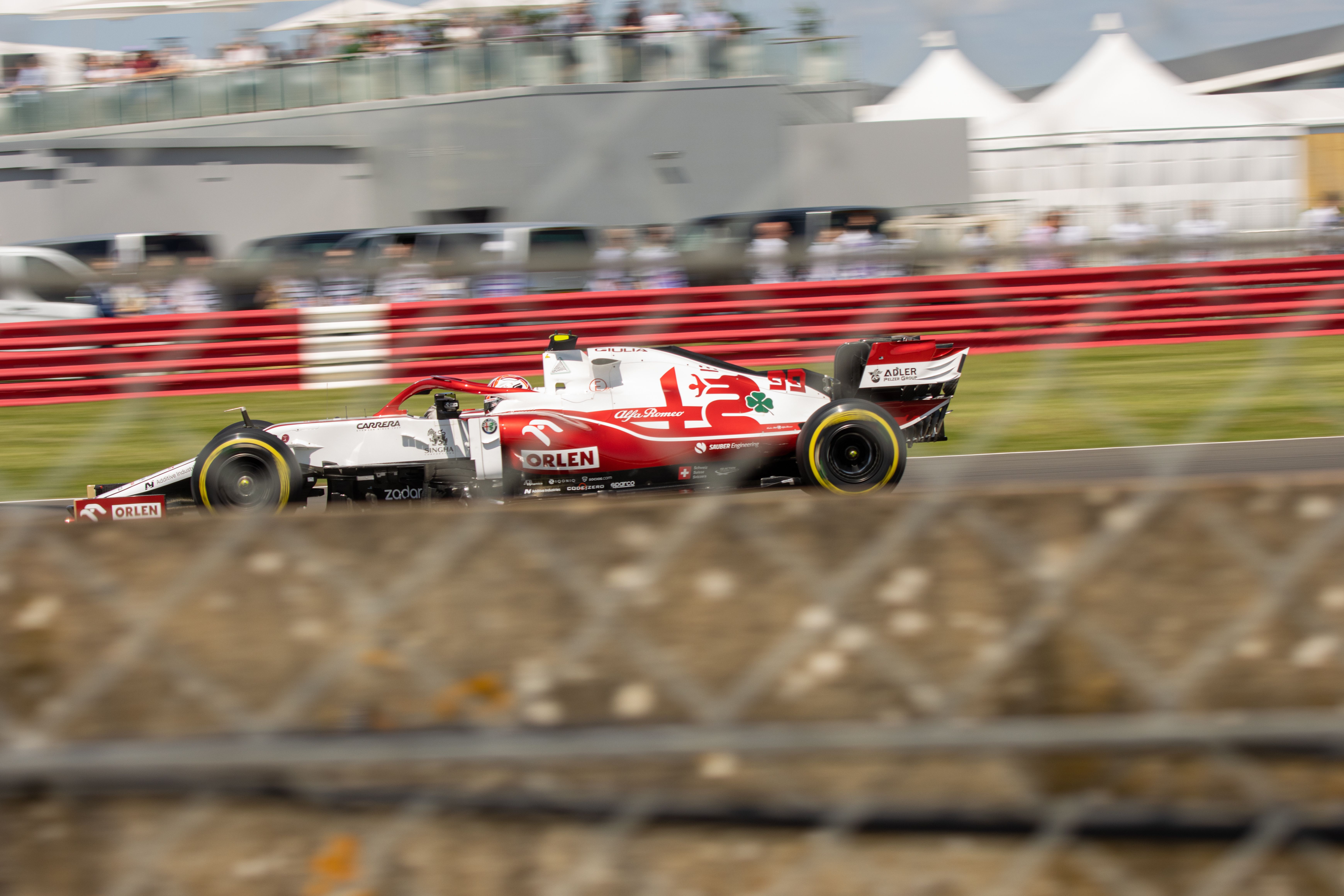 Alfa Romeo Formula One driver Antonio Giovinazzi drives past the camera at high speed, the background is blurred, and the foreground has some chain link fence in soft focus. The subject is the driver in his F1 car. His car is red and white with the number 99 on the side.