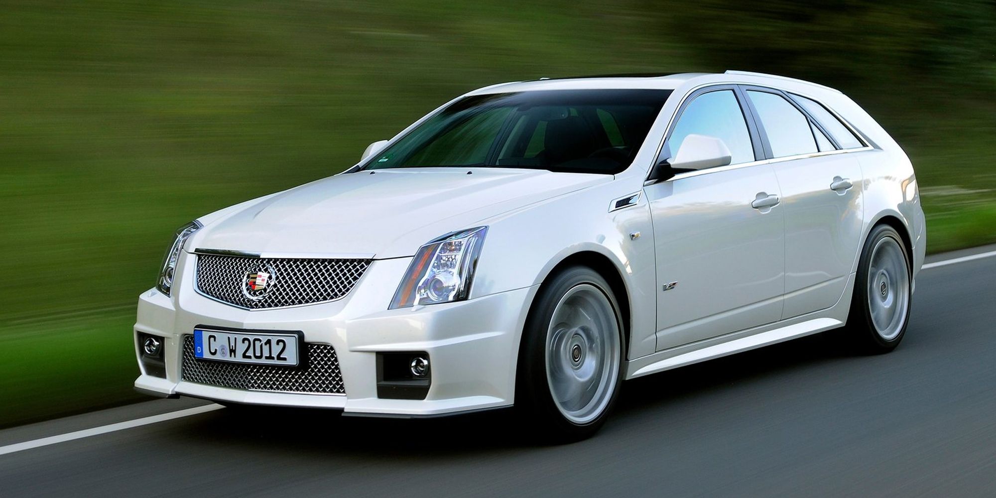 Front 3/4 view of a white CTS-V Sport Wagon on the move, next to a grassy incline