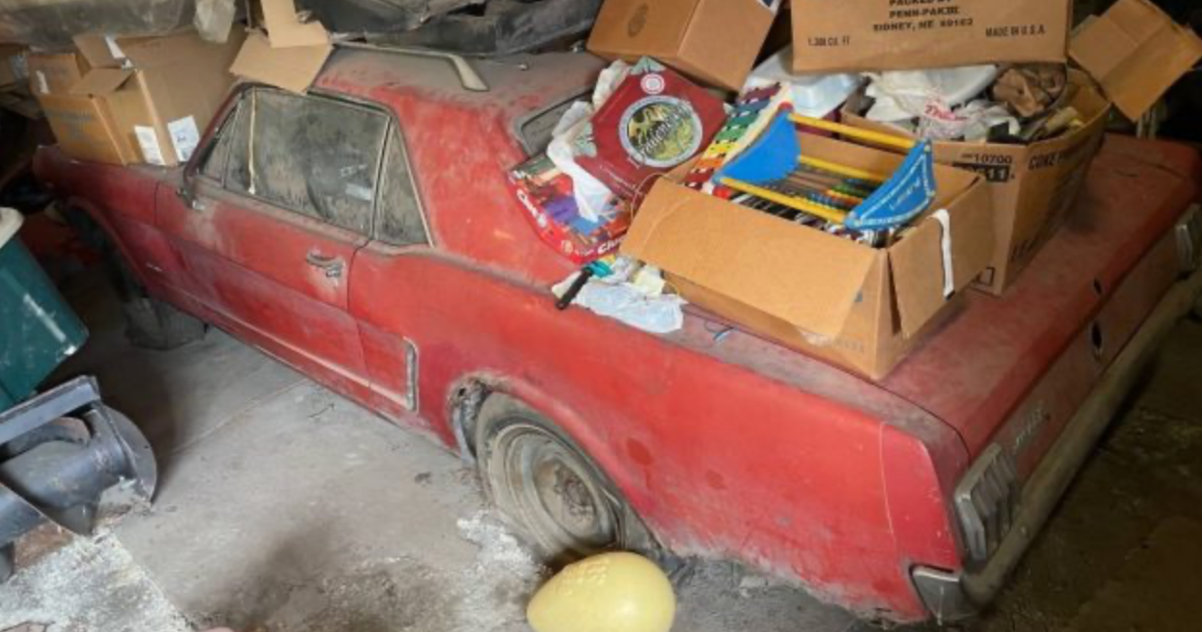 An abandoned classic Red Ford Mustang sits in a garage covered in boxes and dusted